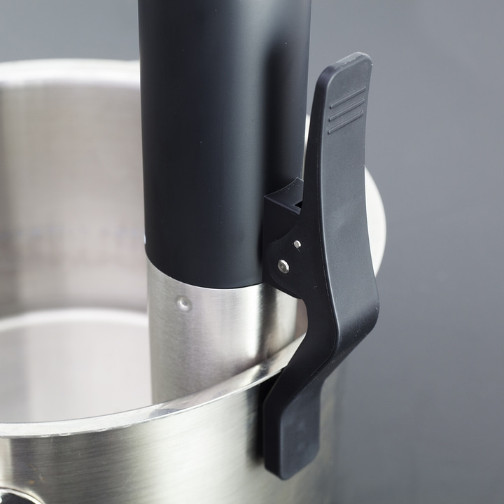 Black device with clamp on metal pot, close-up shot, gray background.