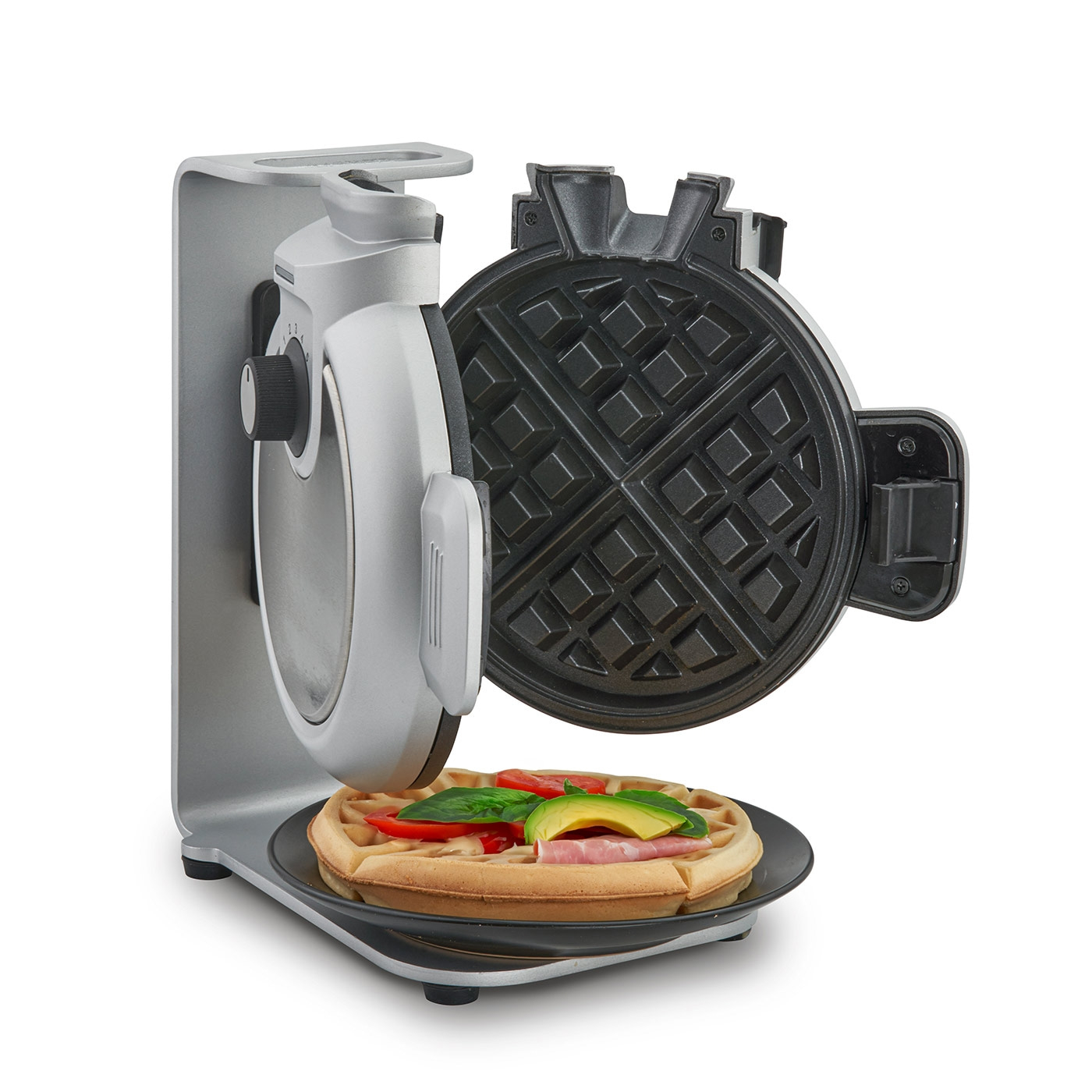 Waffle iron with waffle topped with tomatoes, avocado, and ham on a plate.
