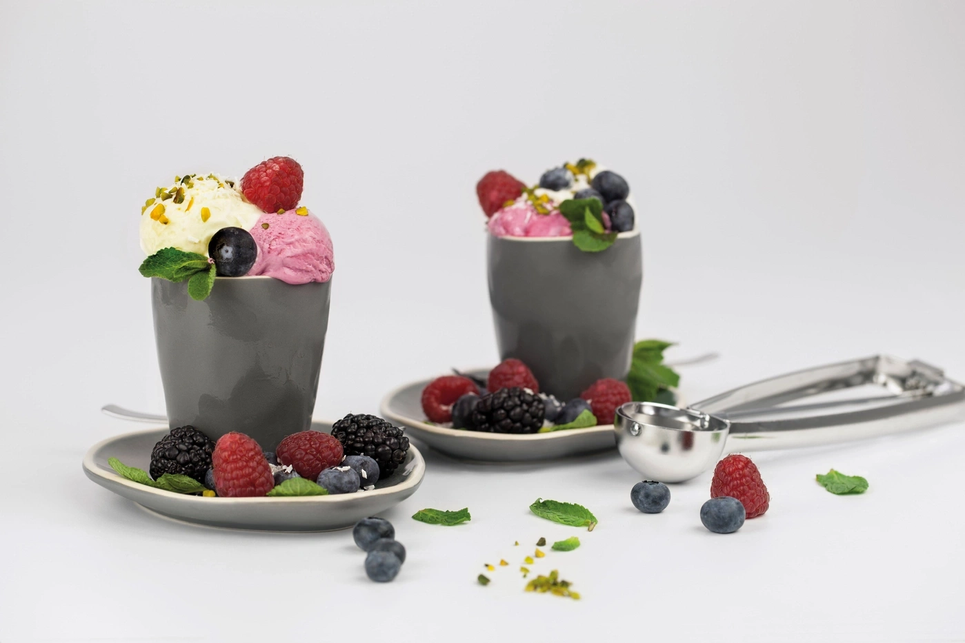 Two ice cream cups with berries and pistachios on saucers, next to an ice cream scoop.
