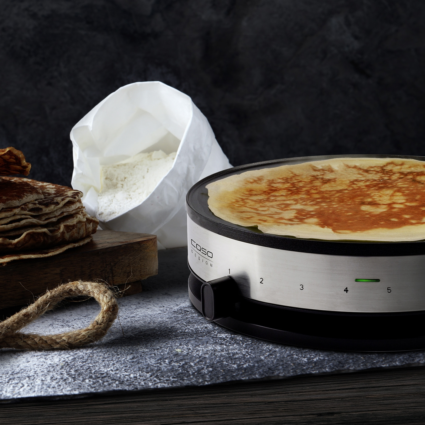 Crêpes-Maker mit Crêpe, Mehl, Holzbrett und Seil auf dunklem Hintergrund.