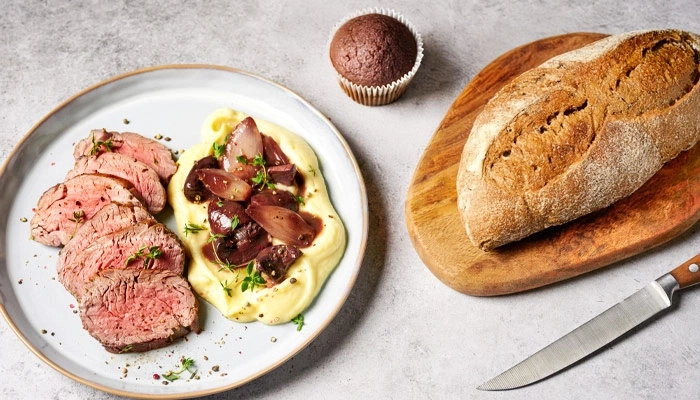 Teller mit Fleisch, Kartoffelpüree, Brot, Kuchen und Messer auf dem Tisch.