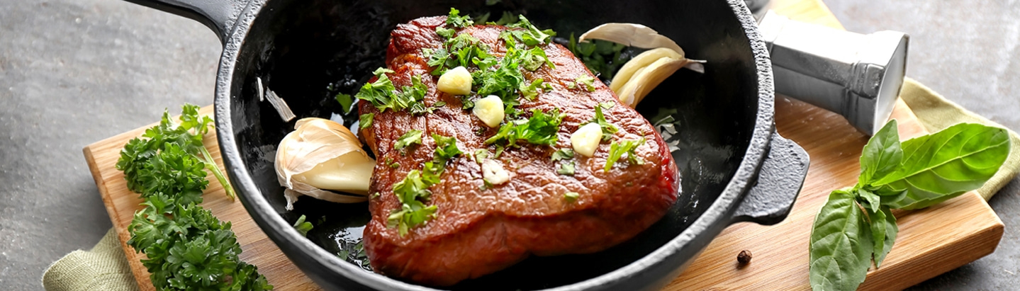 Gebratenes Steak in einer Pfanne mit Knoblauch und Kräutern auf einem Holzbrett.