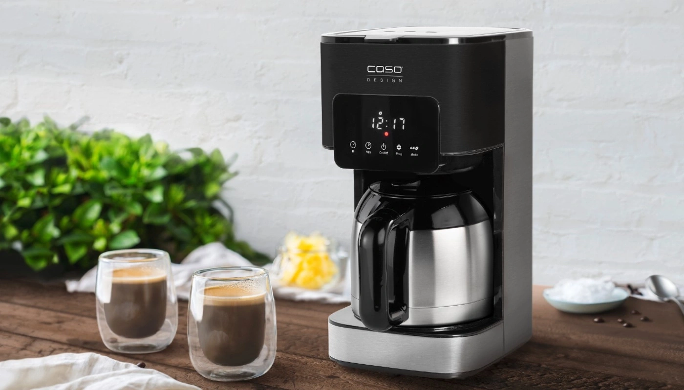 Kaffeemaschine mit zwei Kaffeegläsern auf einem Holztisch, mit Grünzeug im Hintergrund.