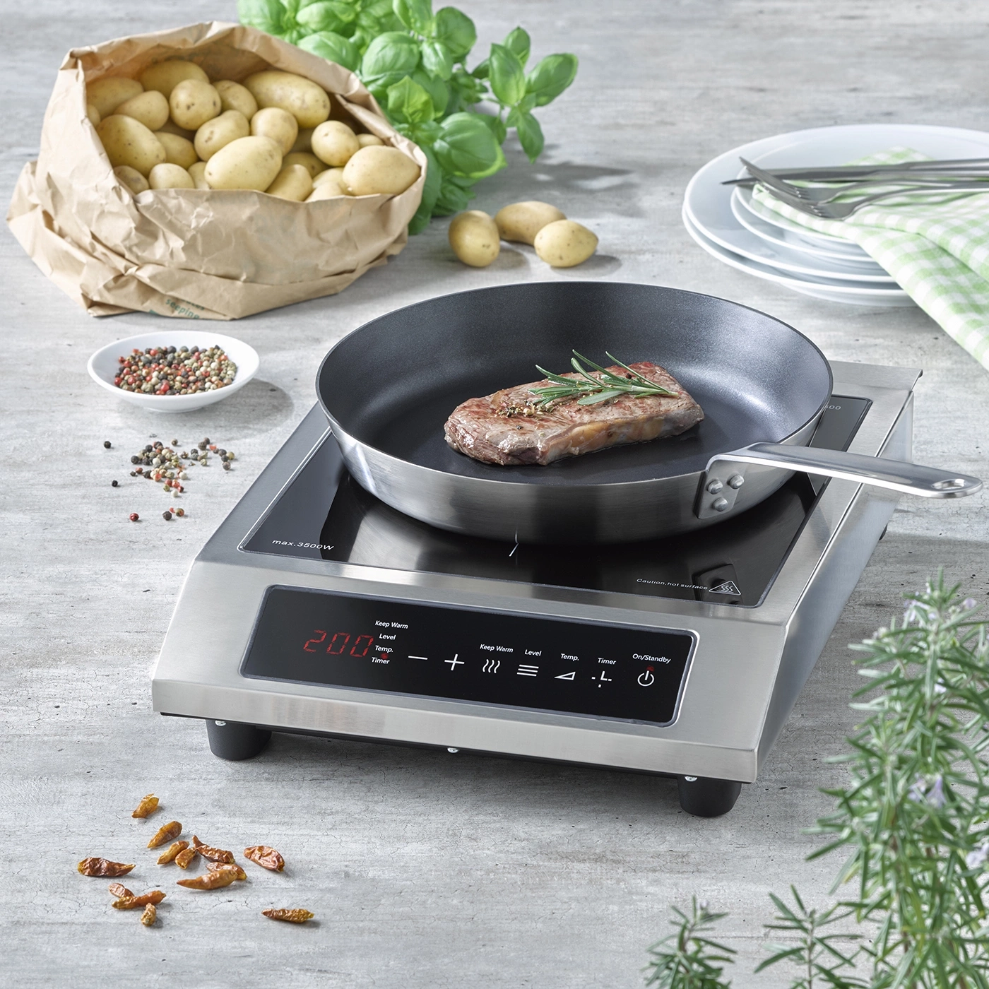 Induction cooktop with steak in pan, potatoes, herbs, and spices on a table setting.