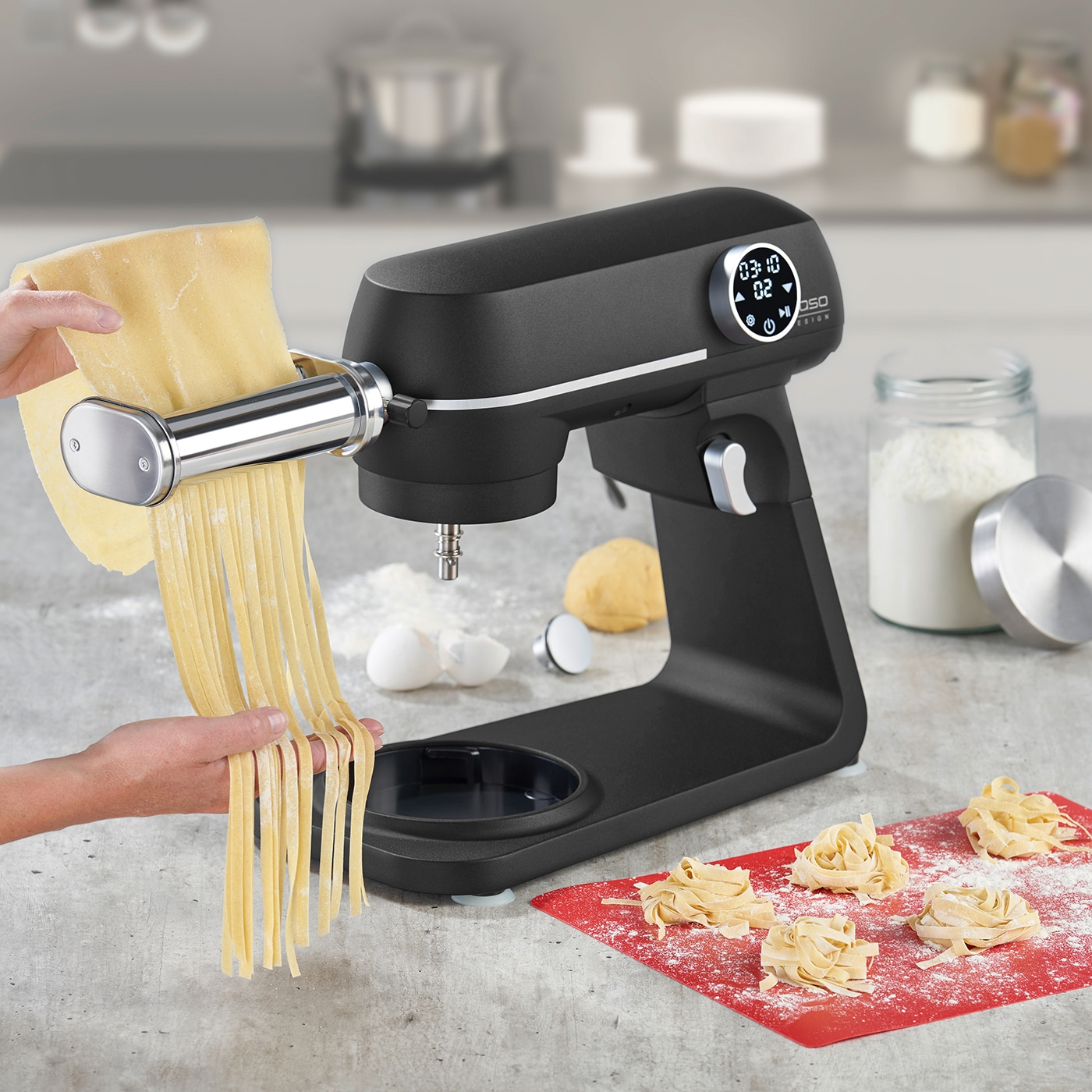 Schwarze Küchenmaschine mit Nudelaufsatz, Nudeln und Zutaten auf dem Tisch.