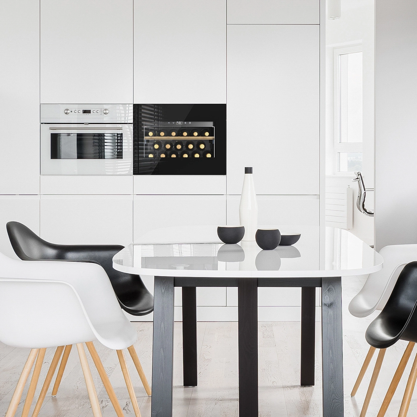 A modern dining area with white and black chairs around a round table in a white kitchen with a wine cooler.