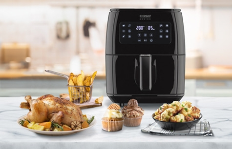 Black air fryer with food including chicken, fries, and muffins on a table.
