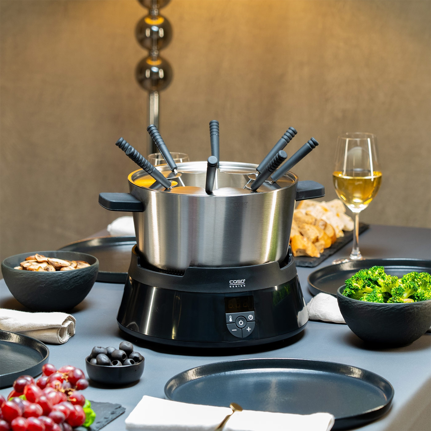 Fondue-Set mit Gemüse, Obst, Brot und Wein auf einem Tisch.