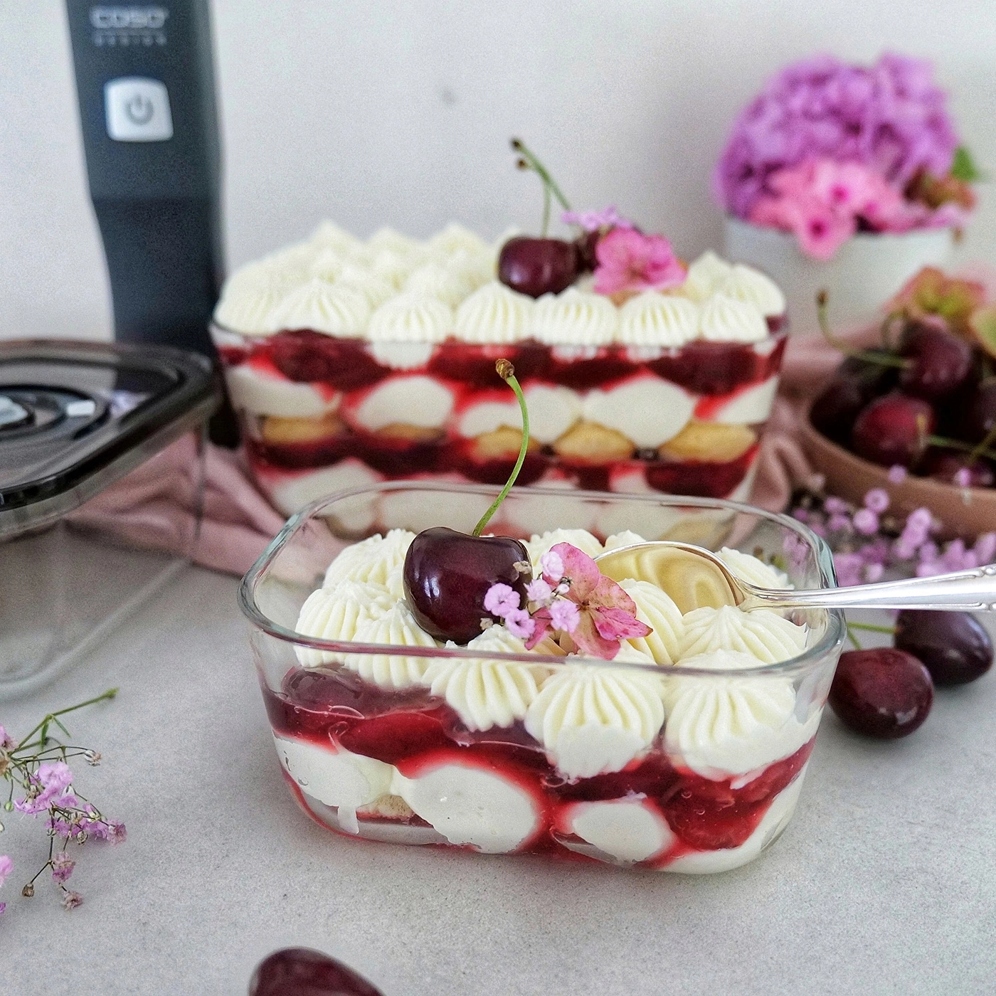 Dessert mit Kirschen und Sahne in Glasbehältern, dekoriert mit Blumen.