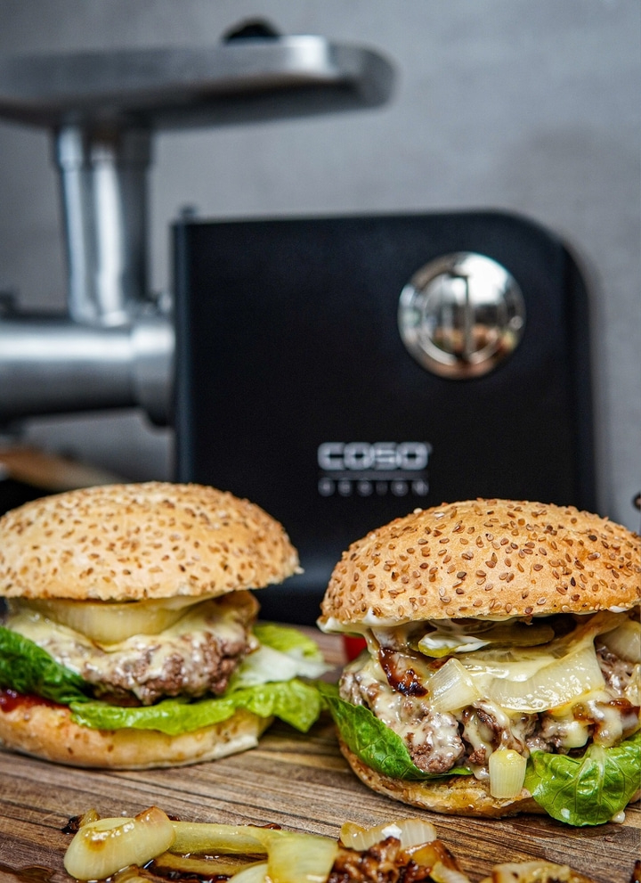 Two burgers on a wooden board with a meat grinder in the background.