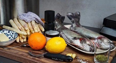Zwei Fische, Spargel, Zitrone, Orange, Butter, Gewürze auf einem Holzbrett.