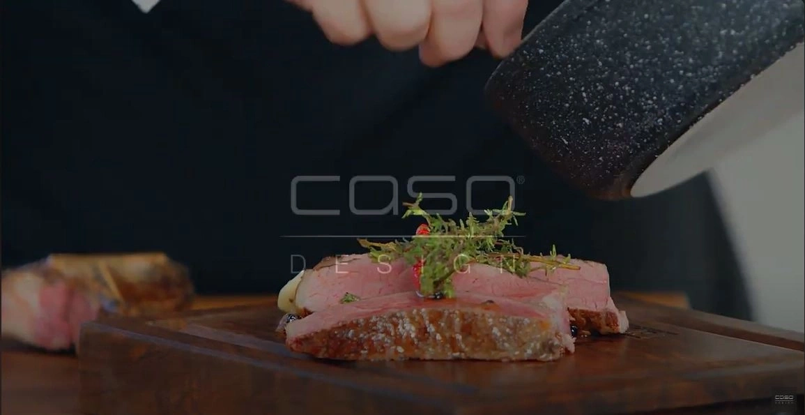 Ein Koch serviert ein Steak mit Kräutern auf einem Holzbrett.