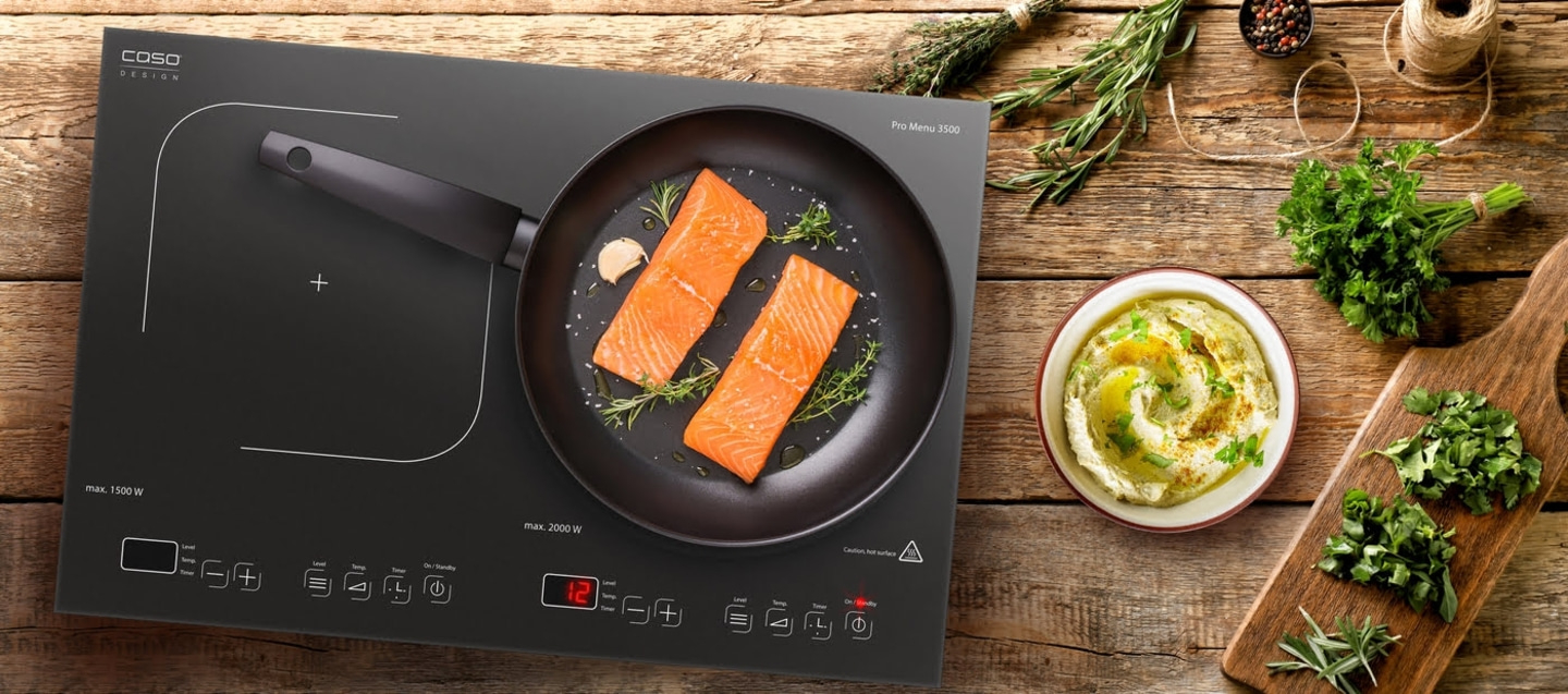 Salmon fillets in a pan on an induction cooktop with herbs and hummus