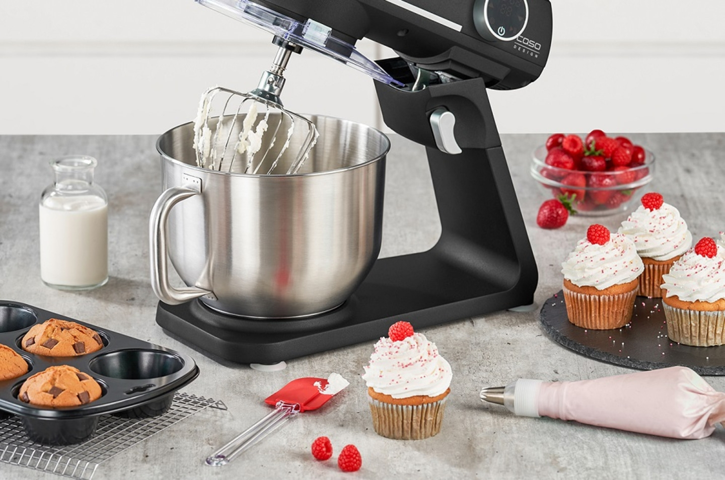 Stand mixer with cupcakes, strawberries, and baking ingredients on a table.