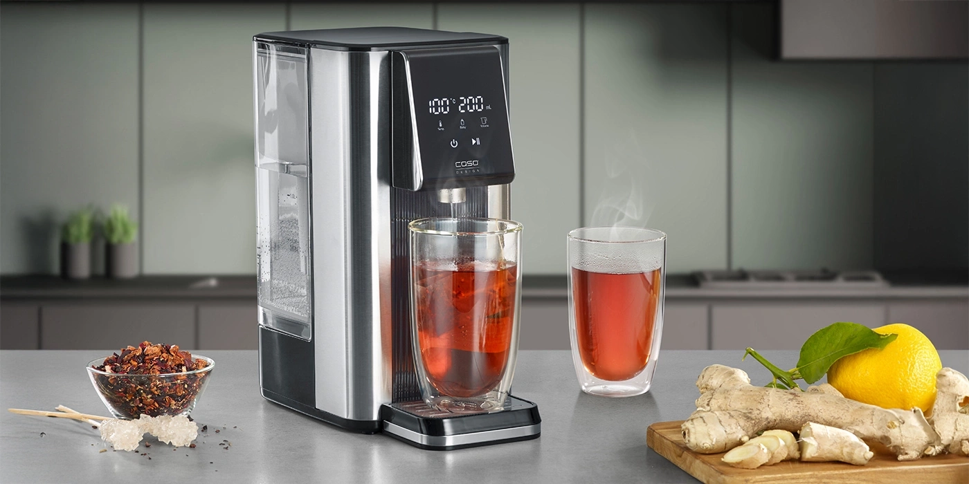 Tea machine with two tea glasses, ginger, lemon, and tea leaves on a table.