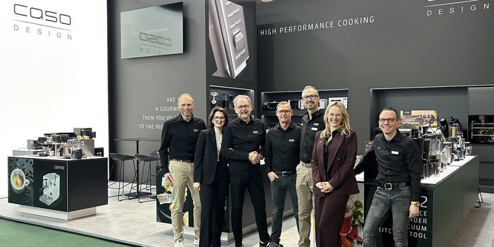 A group of seven people stand in front of a CASO DESIGN exhibition stand with kitchen appliances.