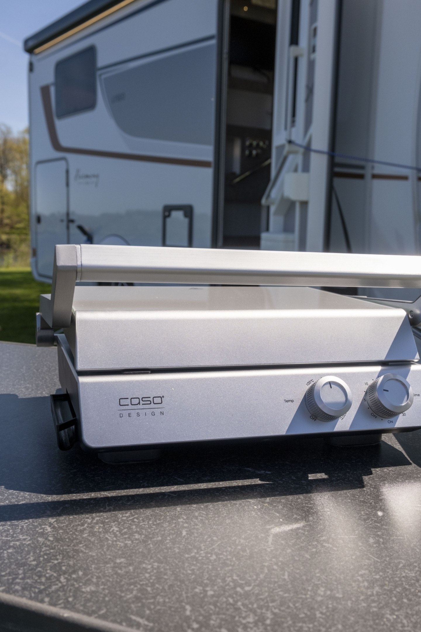 Silberner Kontaktgrill der Marke CASO DESIGN steht auf einem Tisch vor einem Wohnmobil.