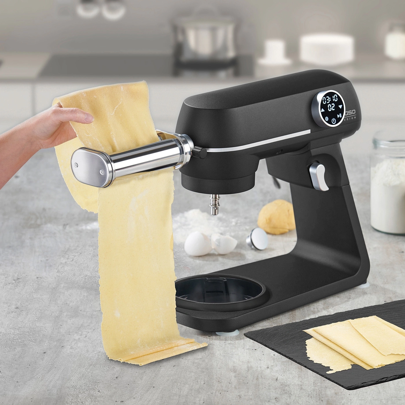 Black kitchen machine with pasta attachment, dough being processed, kitchen in the background.
