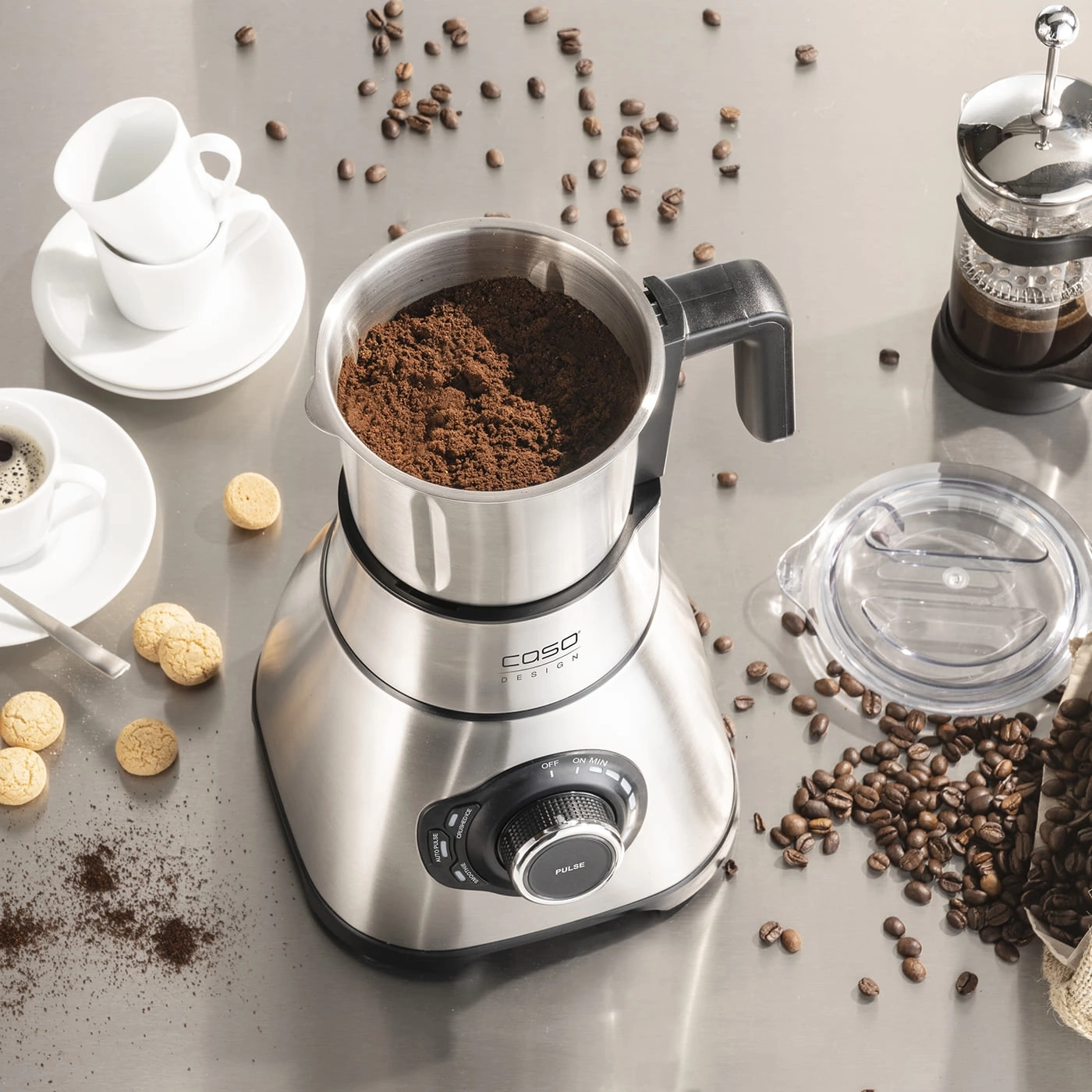 Kaffeemühle mit Kaffeebohnen, Tassen und Gebäck auf einer Metalloberfläche.
