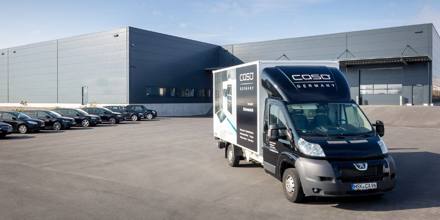 Black delivery van with COSO logo in front of a warehouse with parked cars.