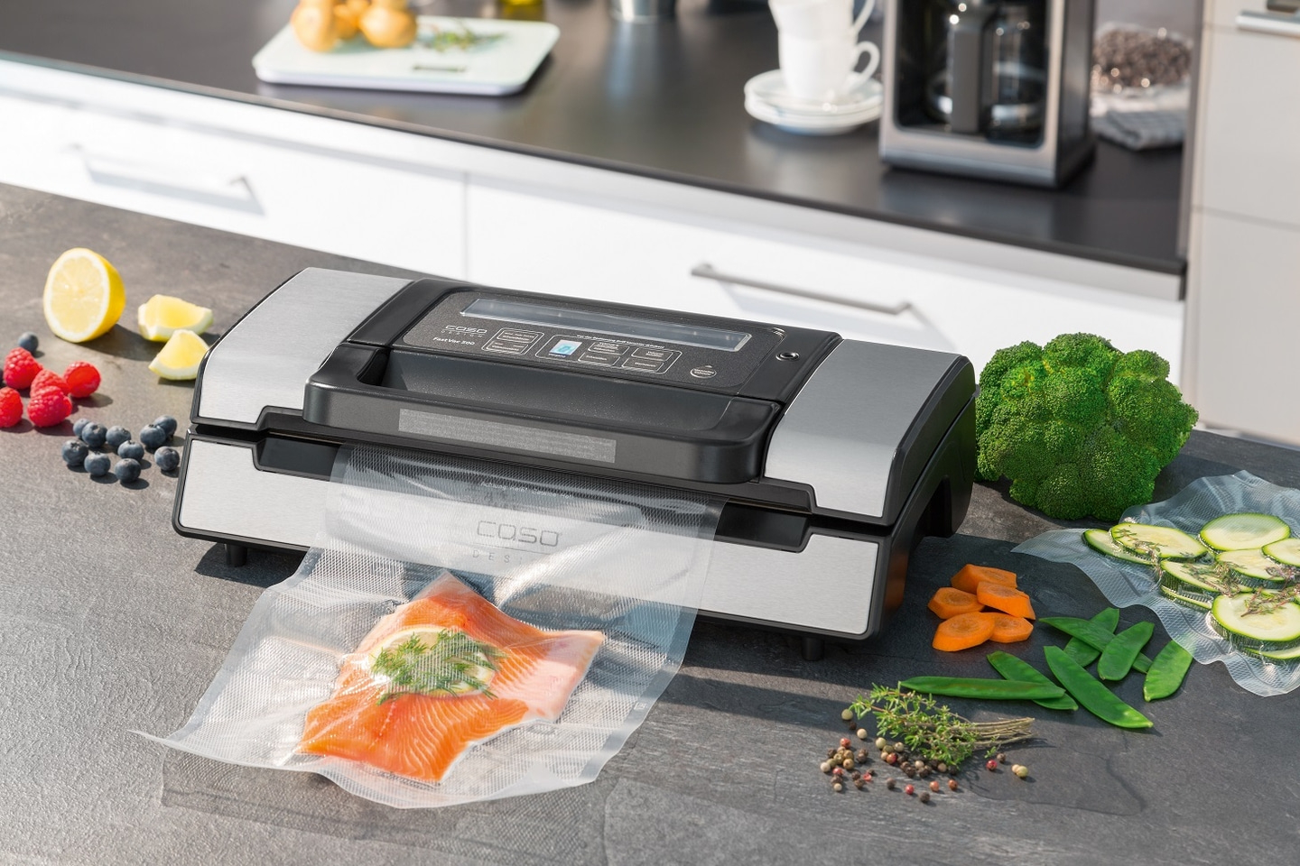 Vakuumiergerät mit Lachs, Gemüse und Früchten auf einer Arbeitsplatte.