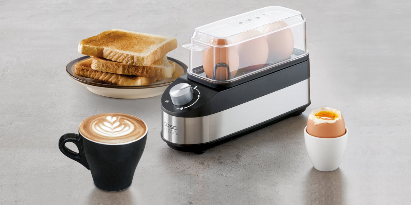 Frühstück mit Toast, Kaffee, Eiern und Eierkocher auf grauem Hintergrund.