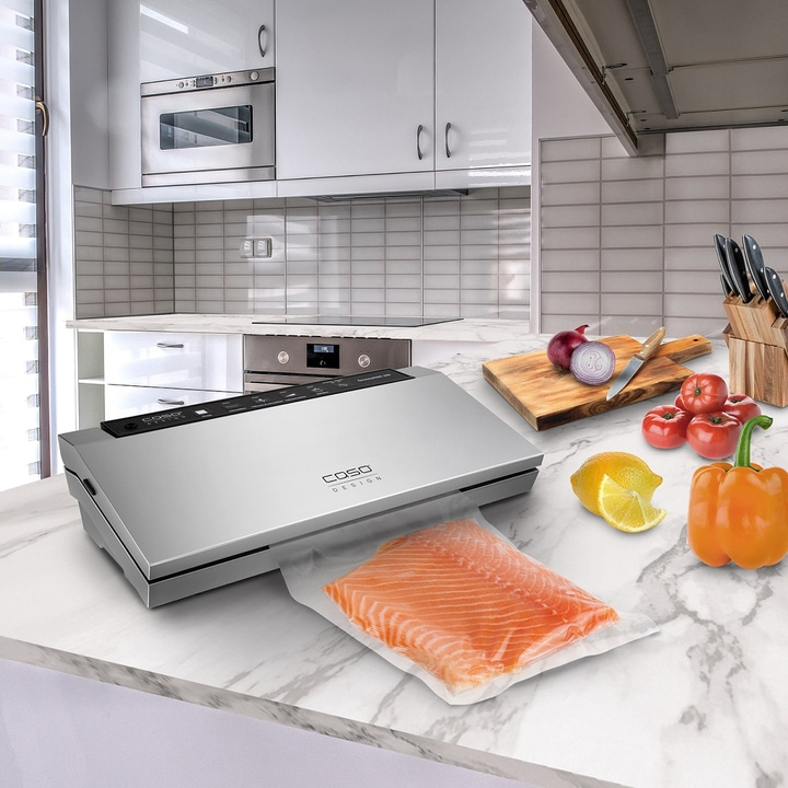 Küche mit Vakuumierer, Lachs, Gemüse und Küchenutensilien auf Marmorplatte.