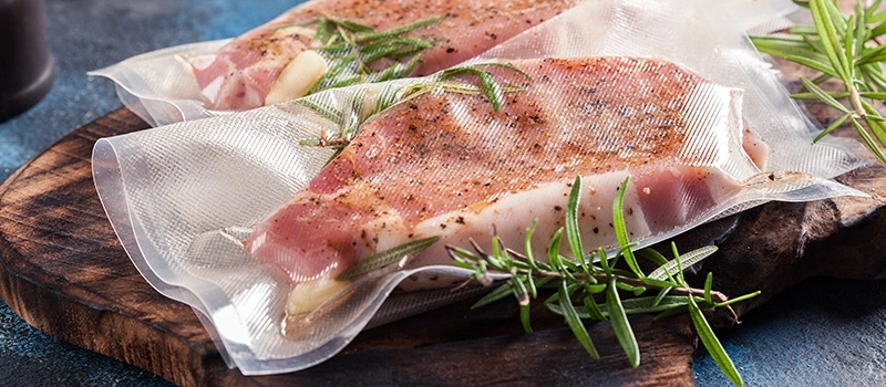 Two vacuum-packed meat pieces with rosemary on a wooden board.