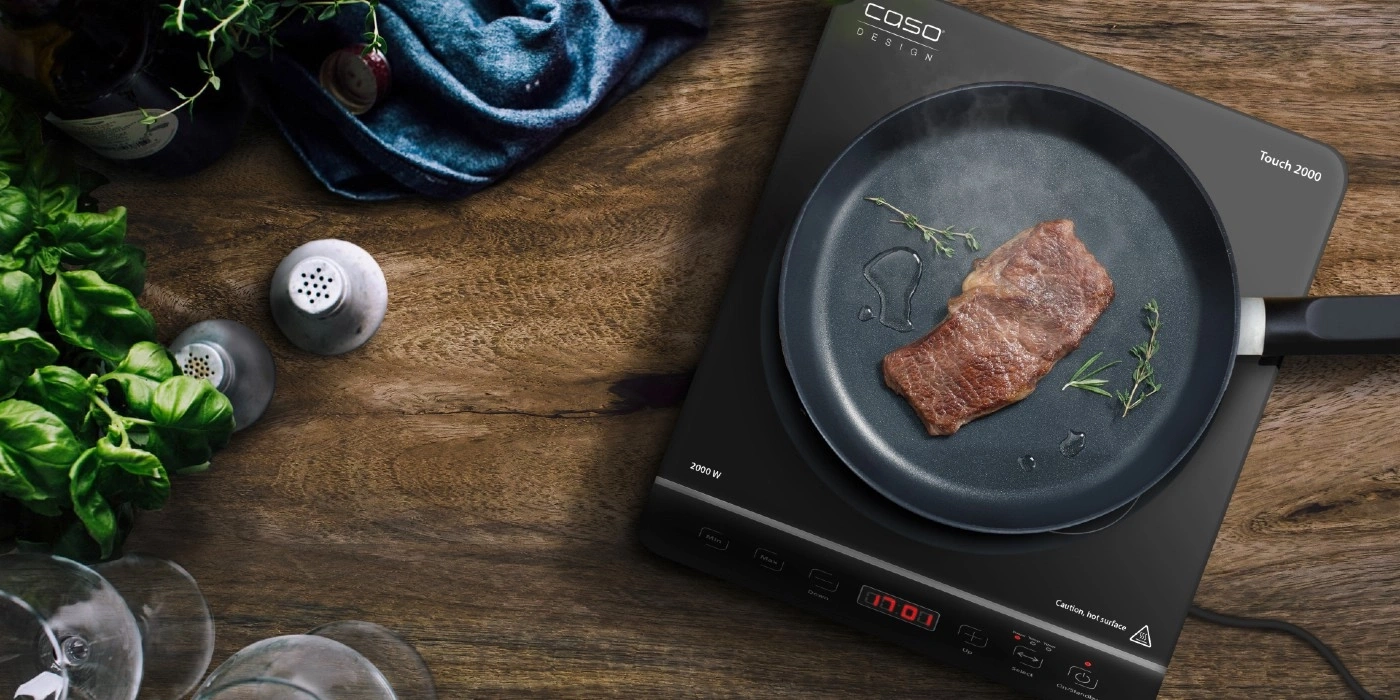 A steak is being cooked on an induction cooktop, surrounded by herbs and ingredients.