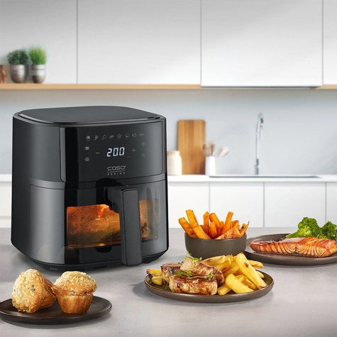 Black air fryer with food on a table in a kitchen setting.