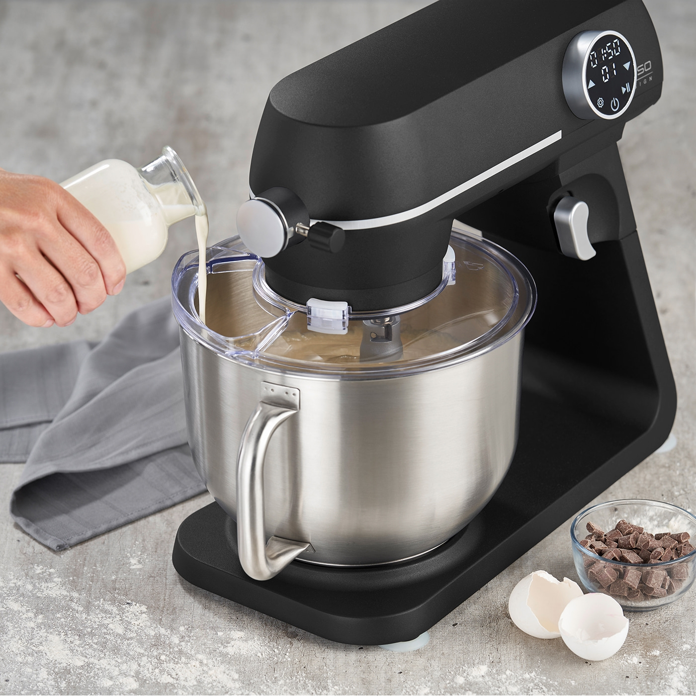 Stand mixer with ingredients, milk being poured, chocolate and eggshells in the scene.