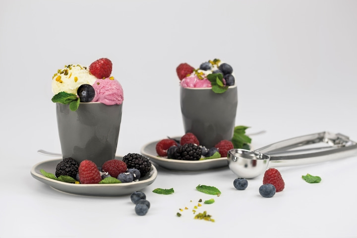 Two ice cream cups with berries and pistachios on saucers, next to an ice cream scoop.