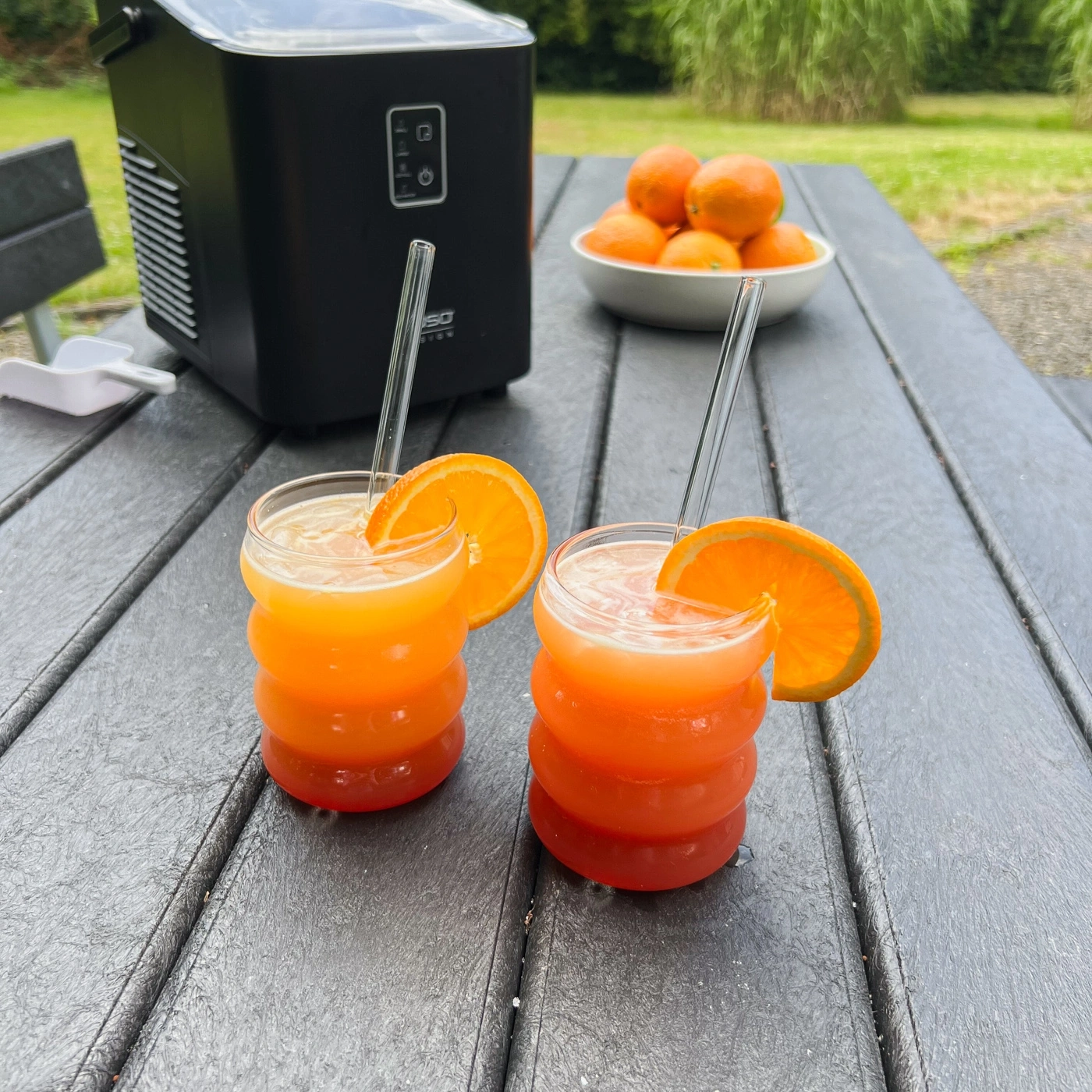 Zwei Cocktails mit Orangenscheiben auf einem Tisch mit Eiswürfelmaschine und Orangen.