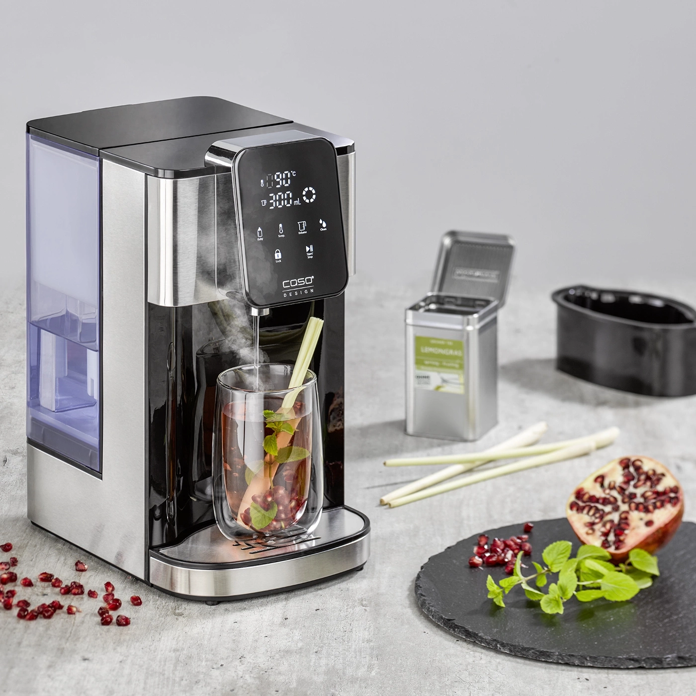 Tea machine with glass, pomegranate, mint, and lemongrass on slate plate.