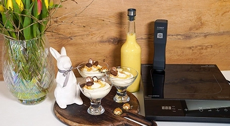 Oster-Dessert mit Schokoladeneiern, Hase, Blumen und Kochutensilien auf Holzuntergrund