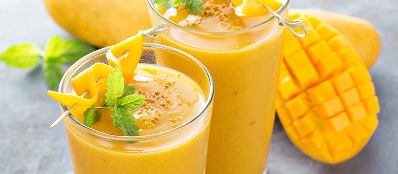 Zwei Gläser Mango-Smoothie mit Minze und Mango-Dekoration, daneben eine Mango.
