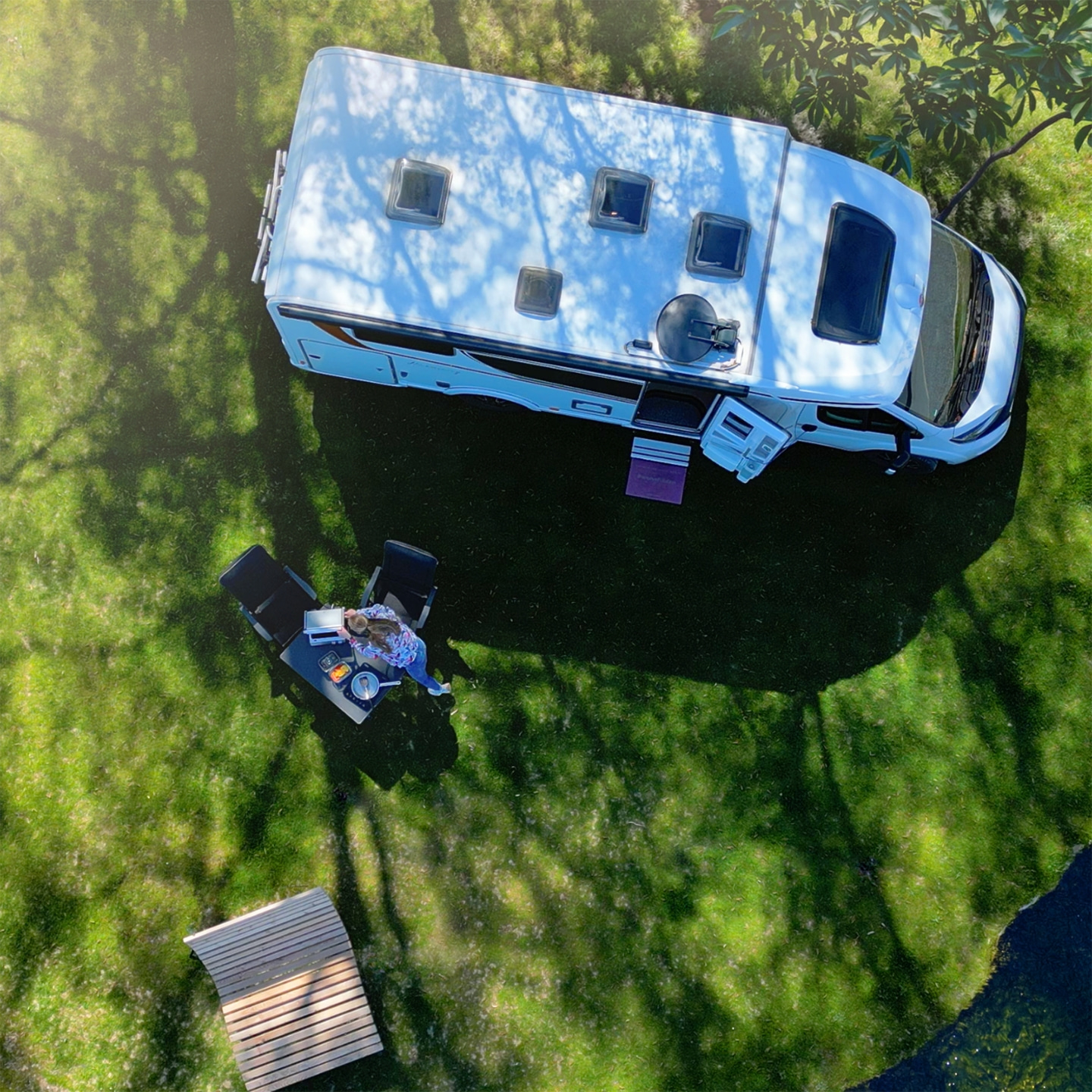 A woman sits at a table in the grass next to a campervan, working on her laptop.