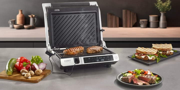 Grill with steaks, vegetables, and sandwiches on a kitchen surface.