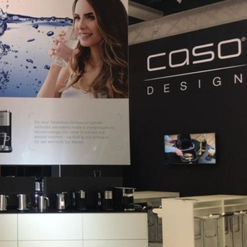 CASO Design stand with woman, water, products. Black wall with logo and a screen.