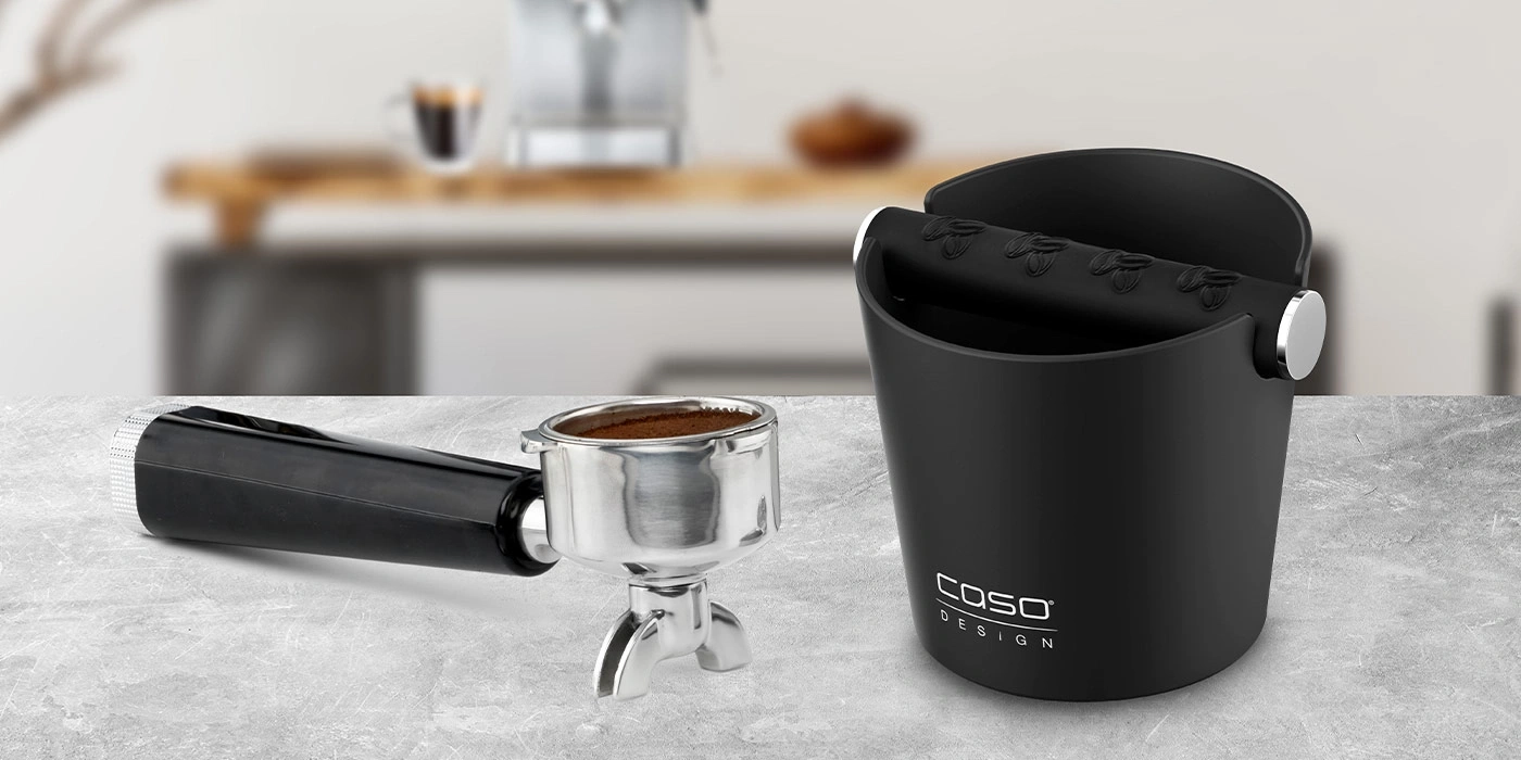 Schwarzer Espresso-Abschlagbehälter und Siebträger mit Kaffeepulver auf grauem Untergrund