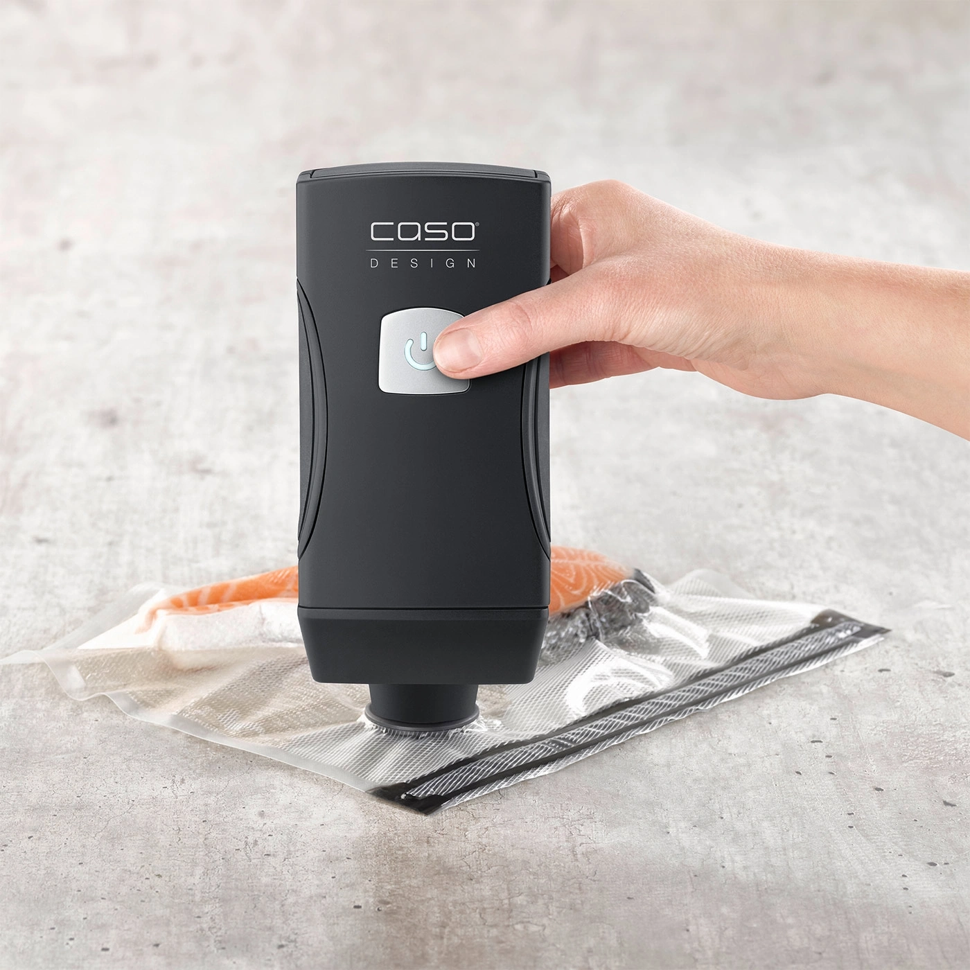 Hand holding vacuum sealer over bag with salmon on gray surface.