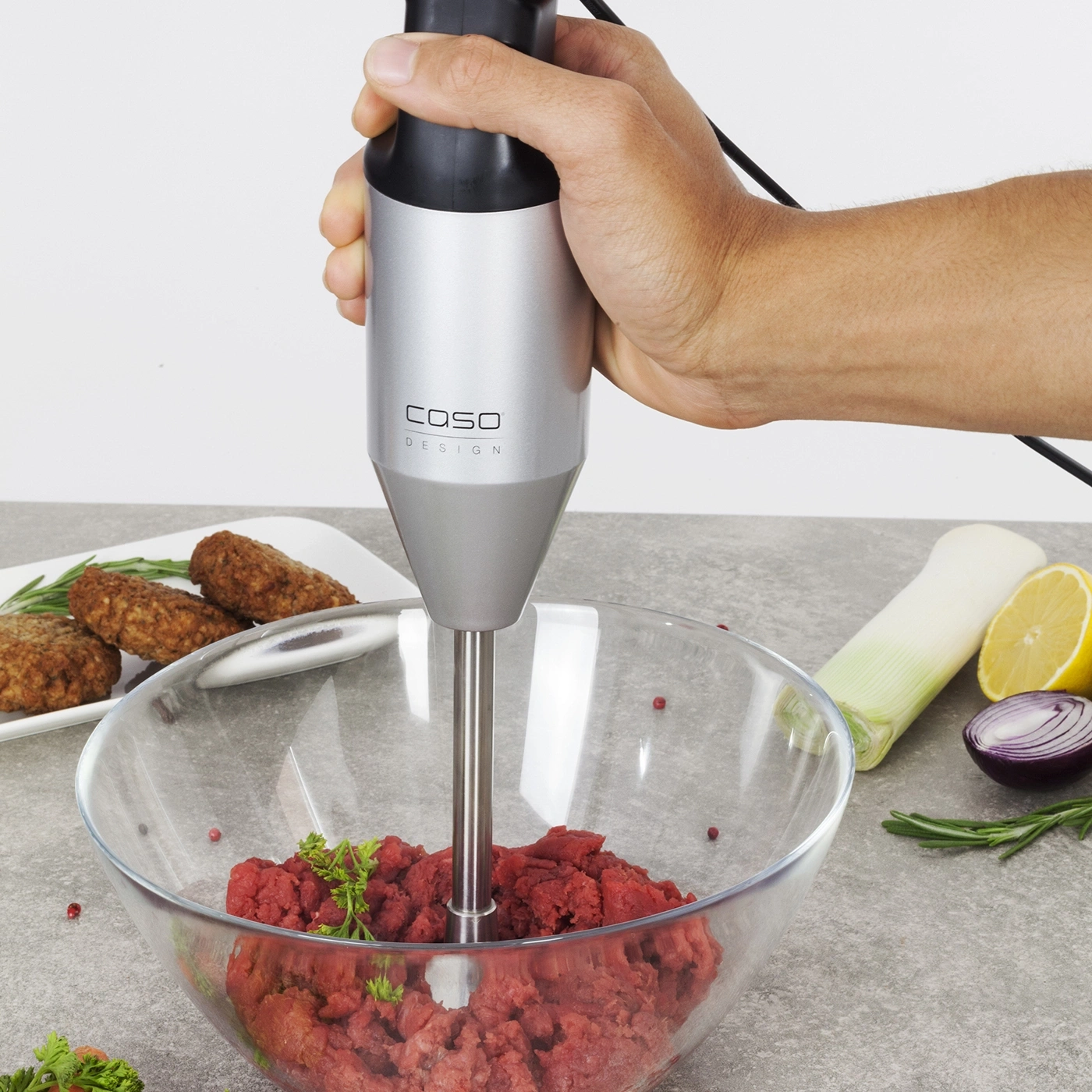 Hand hält Stabmixer über Schüssel mit Hackfleisch, daneben Gemüse und Fleisch