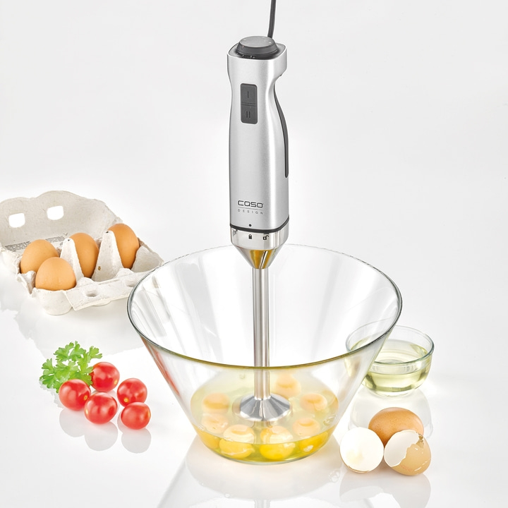 Hand blender in a bowl with eggs, tomatoes, and oil on a white surface.