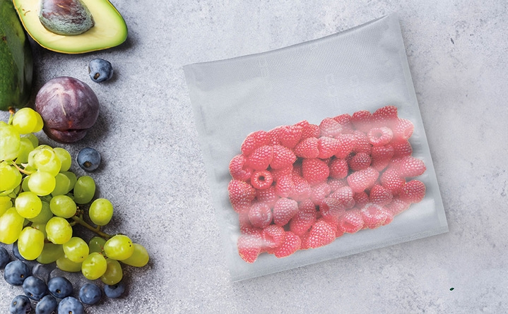 Himbeeren in einer Plastiktüte, daneben Trauben, Avocado und Pflaume auf grauem Untergrund.