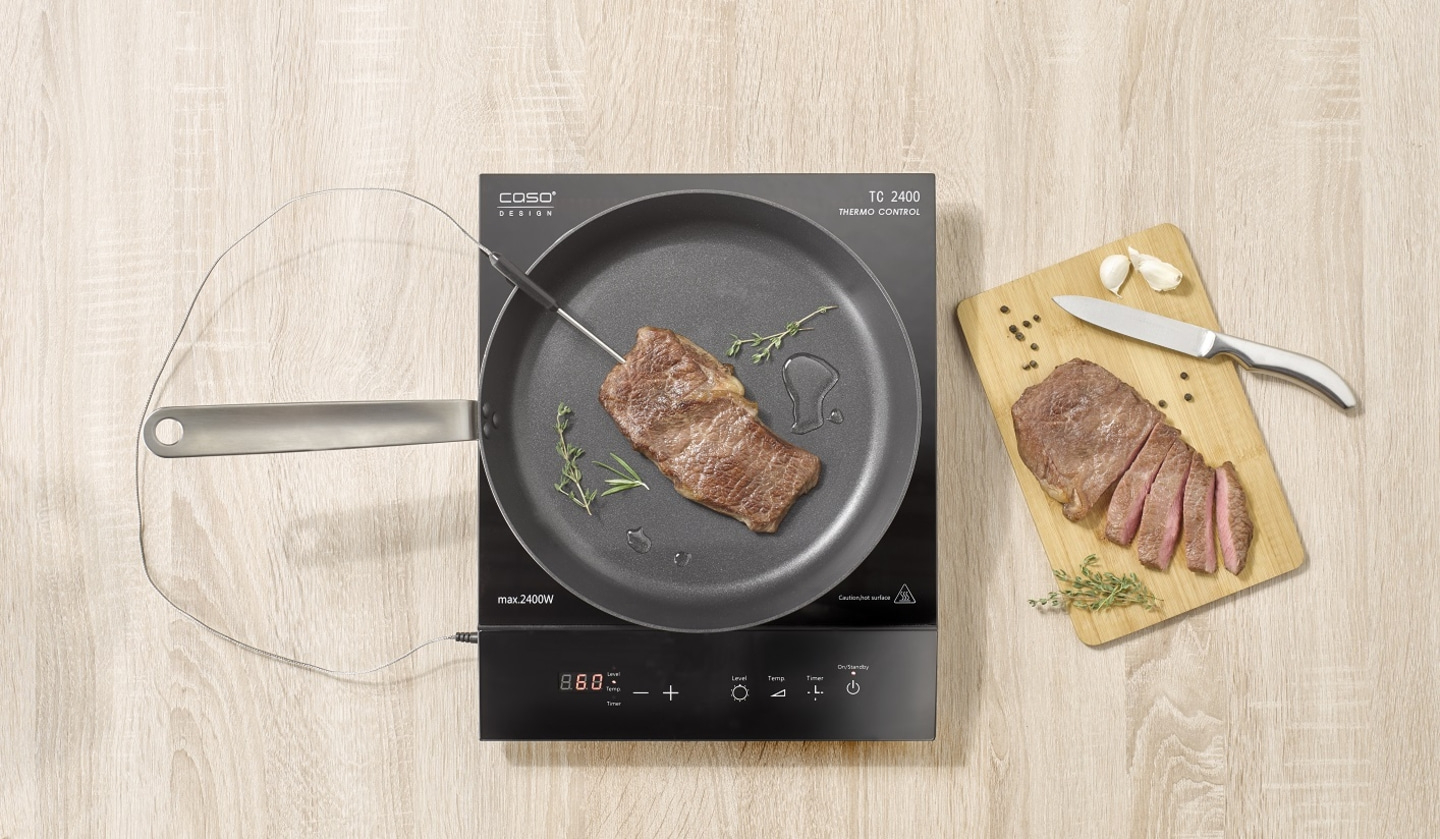 Steak cooking on induction cooktop with thermometer and sliced steak on wooden board