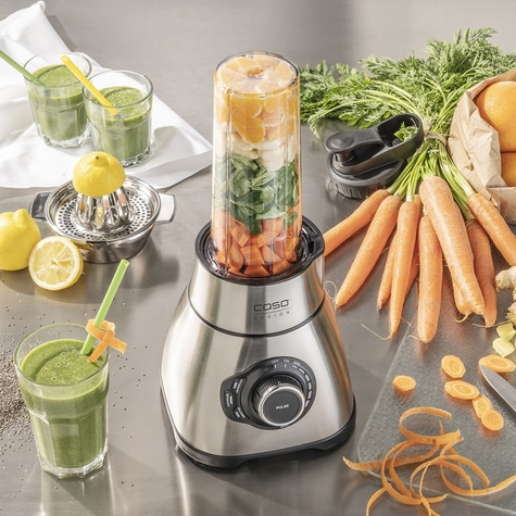 Mixer mit Zutaten, Smoothie-Gläsern und Gemüse auf einer Arbeitsplatte.