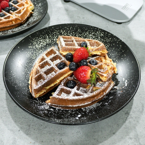 Waffles with strawberries and blueberries on a black plate, dusted with powdered sugar.