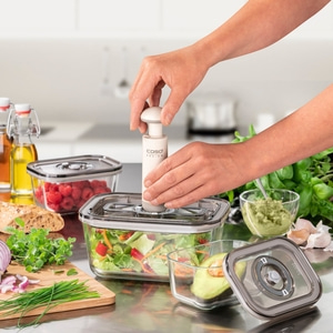 Person using vacuum pump on food containers with vegetables and fruits on a table.