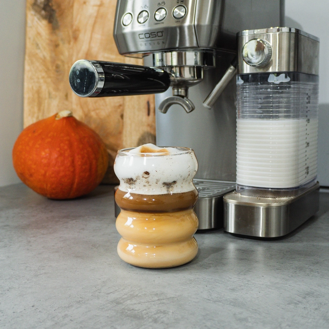 Coffee in a unique glass next to a coffee machine and a pumpkin.