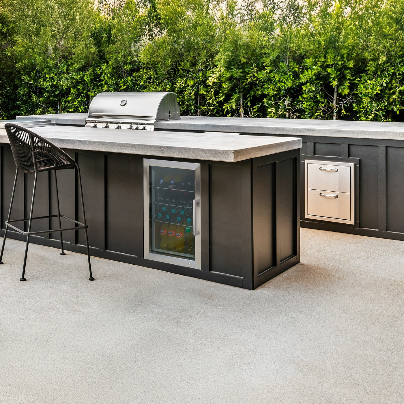Outdoor-Küche mit Grill, Kühlschrank und Barhocker vor grünen Pflanzen.